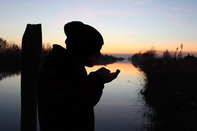 Side view of silhouette woman standing by lake against sky during sunset