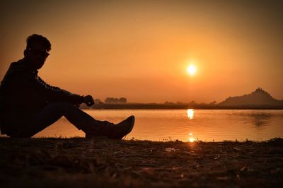 Man sitting at river bank against sky during sunset