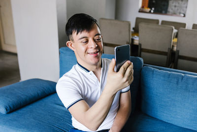 Smiling teenage latin boy with down syndrome taking self shot on smartphone while sitting on sofa at home