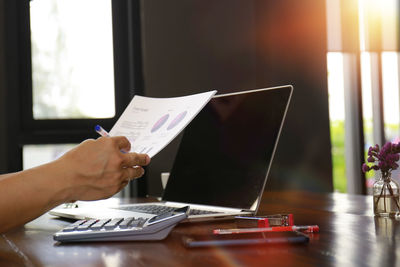 Person holding pen while sitting with laptop at desk in office