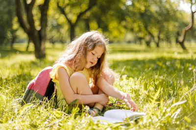 A cute schoolgirl is sitting on the grass in the park and reading a book