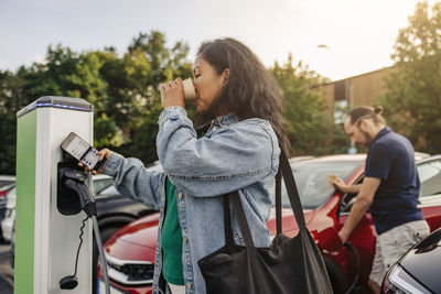 Woman having drink while operating kiosk at charging station