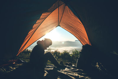 Side view of silhouette man sitting in tent on mountain during sunrise