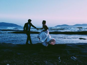 Silhouette couple standing at beach against sky during sunset