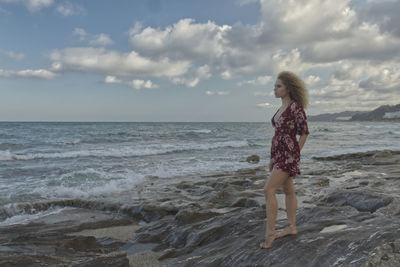 Full length of woman looking at sea while standing on rocky coastline against sky