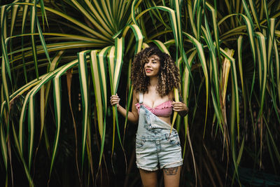 Seductive woman with curly hair standing against plants
