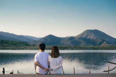 Rear view of couple holding hands while sitting by lake against mountains