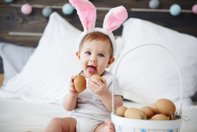 Portrait of cute baby girl with eggs sitting on bed at home