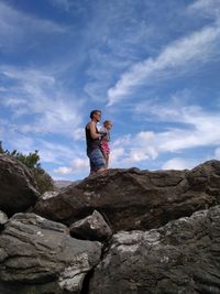 Side view of man with daughter standing on rock against cloudy sky