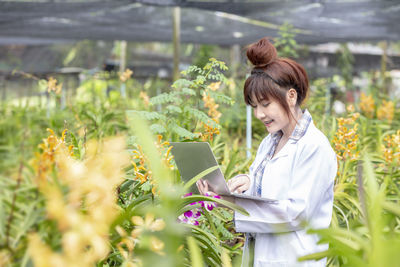 Smiling scientist using laptop amidst plants at greenhouse