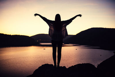 Rear view of woman with arms outstretched on rock by lake against sky during sunset