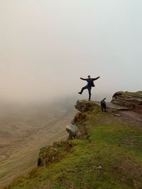 Man with arms outstretched standing on mountain against sky during foggy weather