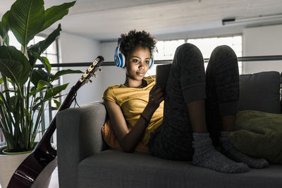 Young woman on couch with headphones using tablet next to guitar