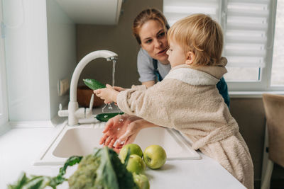 In the bright kitchen, mother and little daughter wash vegetables to prepare dinner together. 