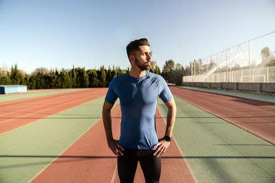 Male sportsperson with hands on hip standing on sports track against sky during sunny day