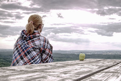 Young blond girl trekker resting at wooden table and taking break from hike. green apple on table