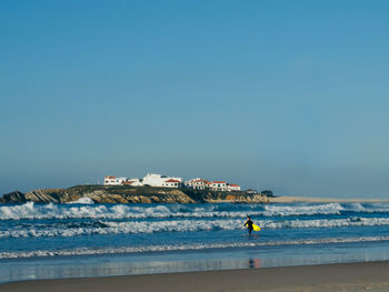 Mid distance view of man with surfboard walking in sea against clear sky