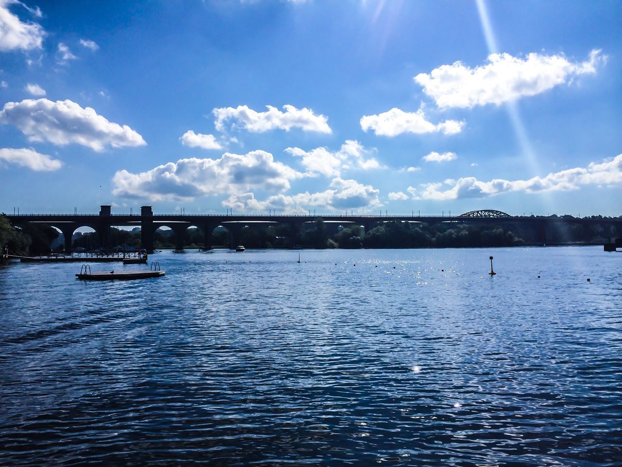 water, bridge - man made structure, waterfront, connection, sky, built structure, river, architecture, rippled, cloud - sky, bridge, sunlight, transportation, tranquility, engineering, blue, tranquil scene, cloud, nature, sunbeam