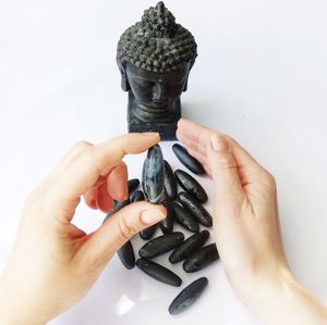 Close-up of hand holding black pebble by buddha statue over white background