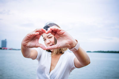 Portrait of woman making heart shape with hands against sea and sky in city