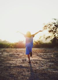 Rear view of girl standing with arms outstretched on field against bright sky
