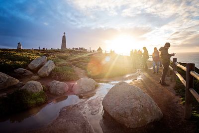 People on rocks by sea against sky during sunset