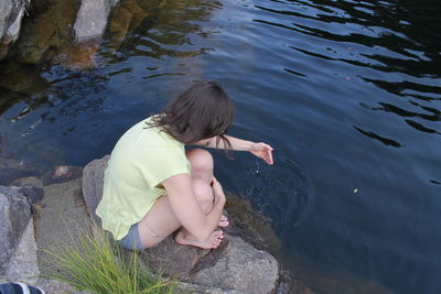 High angle view of woman playing with water while sitting on rock at lakeshore