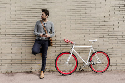 Portrait of young man with racing cycle and cell phone leaning against wall