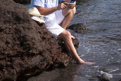 Low section of man using smart phone while sitting on rock by sea