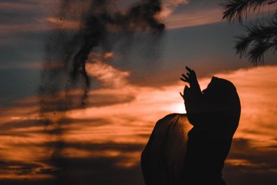 Silhouette woman throwing sand mid air against orange sky during sunset