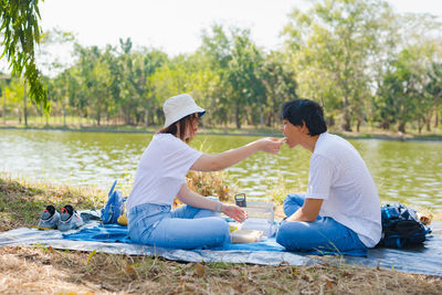 Side view of woman feeding food to boyfriend against lake in park