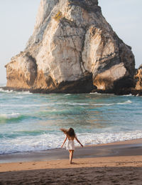 Back view female traveler walking on sandy coast with rough rocky cliffs near sea at sunset time on sunny summer evening