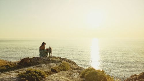 Rear view of woman with dog sitting on rock by sea against sky during sunset