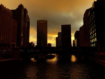 Modern buildings by river against sky during sunset