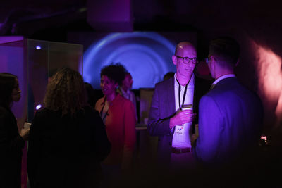 Multiracial male and female colleagues discussing in illuminated exhibition at convention center