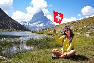 Portrait of smiling woman holding swiss flag while sitting on grassy field by lake