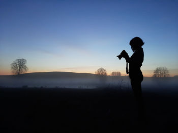 Side view of silhouette woman standing against sky during sunset holding a camera