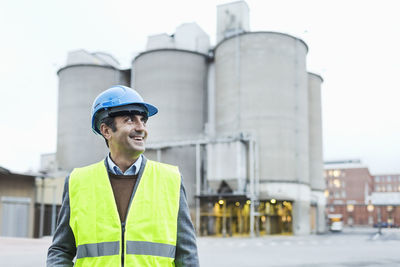 Smiling male manual worker looking away while standing against cement silos