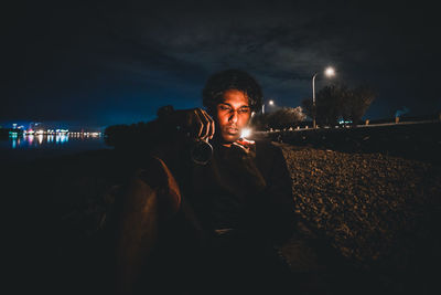 Young man sitting on illuminated street against sky at night