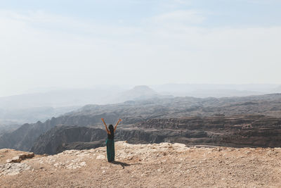 Full length of woman with arms raised standing by cliff against sky