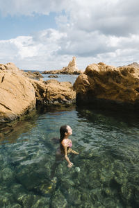 Female tourist in swimwear floating in clean transparent water of sea near rocky rough cliff in summer day
