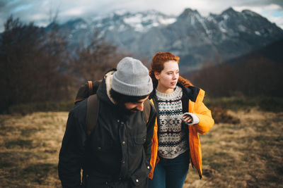 Amorous man with backpack holding hands with beautiful redhead woman in filed with dry grass near mountains in cloudy day