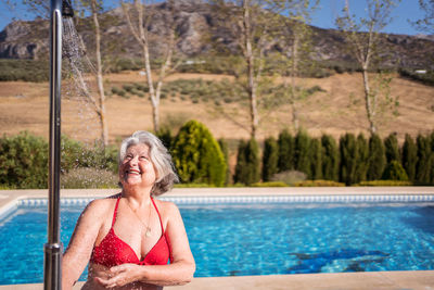 Cheerful senior female in bikini enjoying splashes from shower near pool with transparent clear water