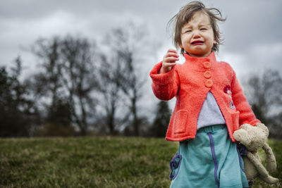 Low angle view of baby girl crying while standing on grassy field against cloudy sky at park