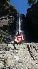 Low angle view of woman meditating on rock against waterfall