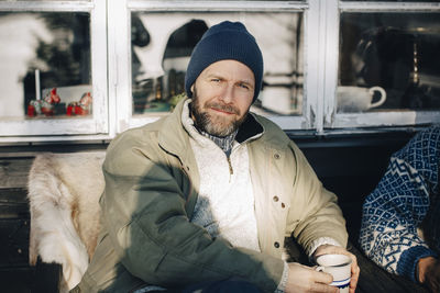 Portrait of smiling mature man sitting with coffee cup at porch in winter