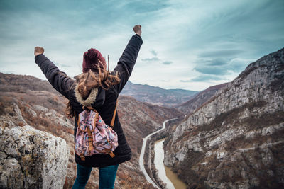 Rear view of woman with arms raised looking at mountains against sky