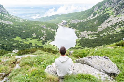 Rear view of man sitting on rock against river and mountains