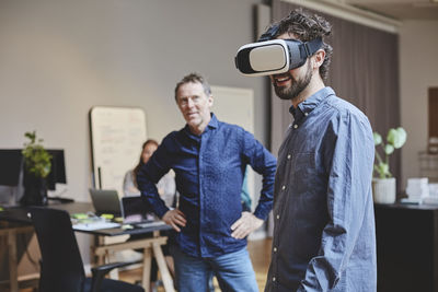 Mature businessman looking at male colleague using virtual reality simulator while standing in creative office