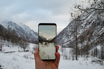 Cropped hand of person photographing snow covered landscape on mobile phone